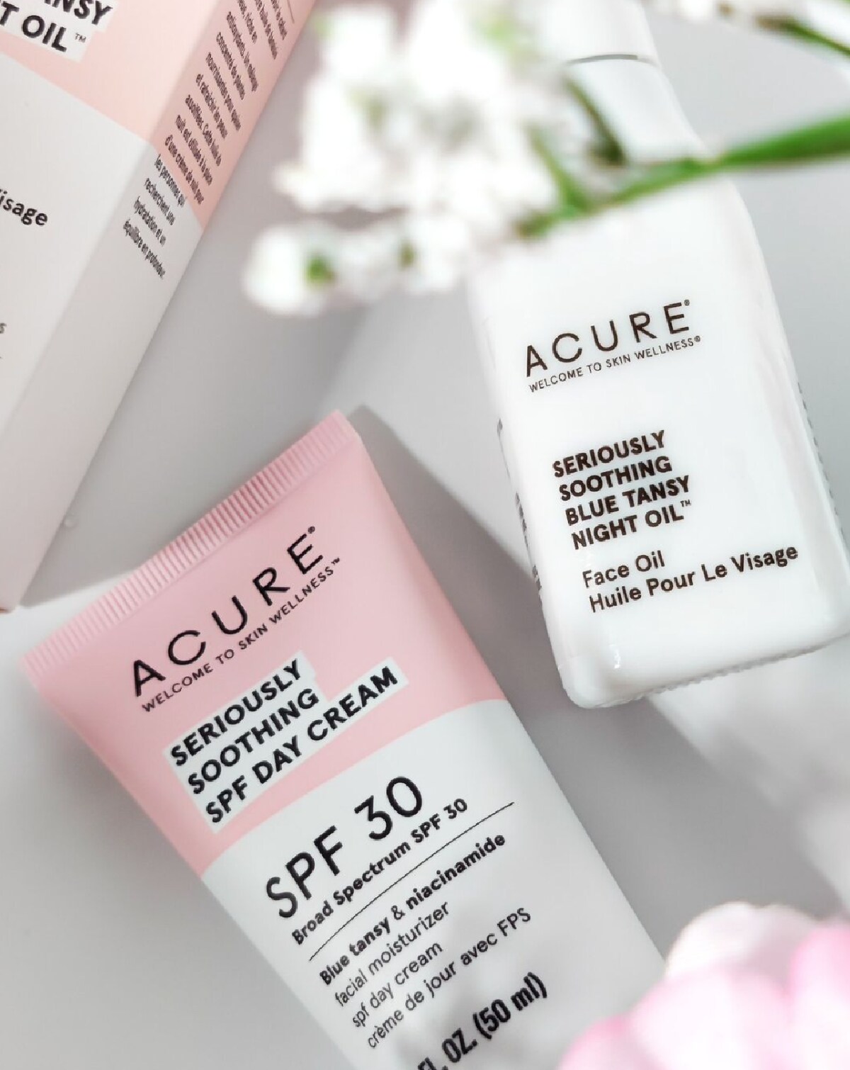 Acure Beauty's white and pink tube of day cream and white bottle of face oil on a white background with white and pink flowers.
