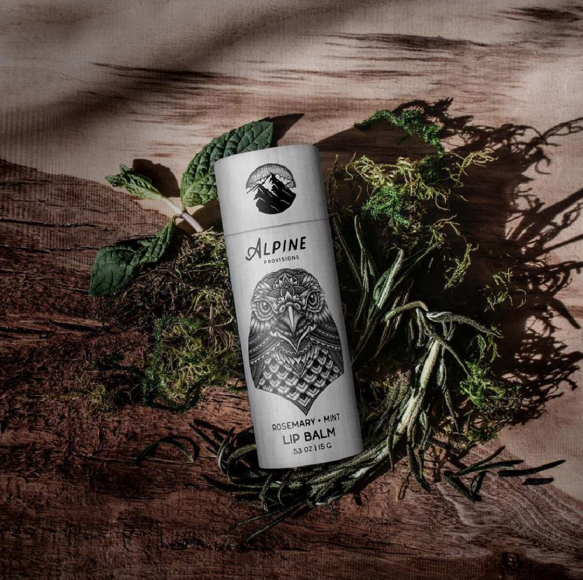 A gray and black Alpine Provisions cardboard tube of vegan lip balm on top of branches of rosemary and mint on a wooden background.