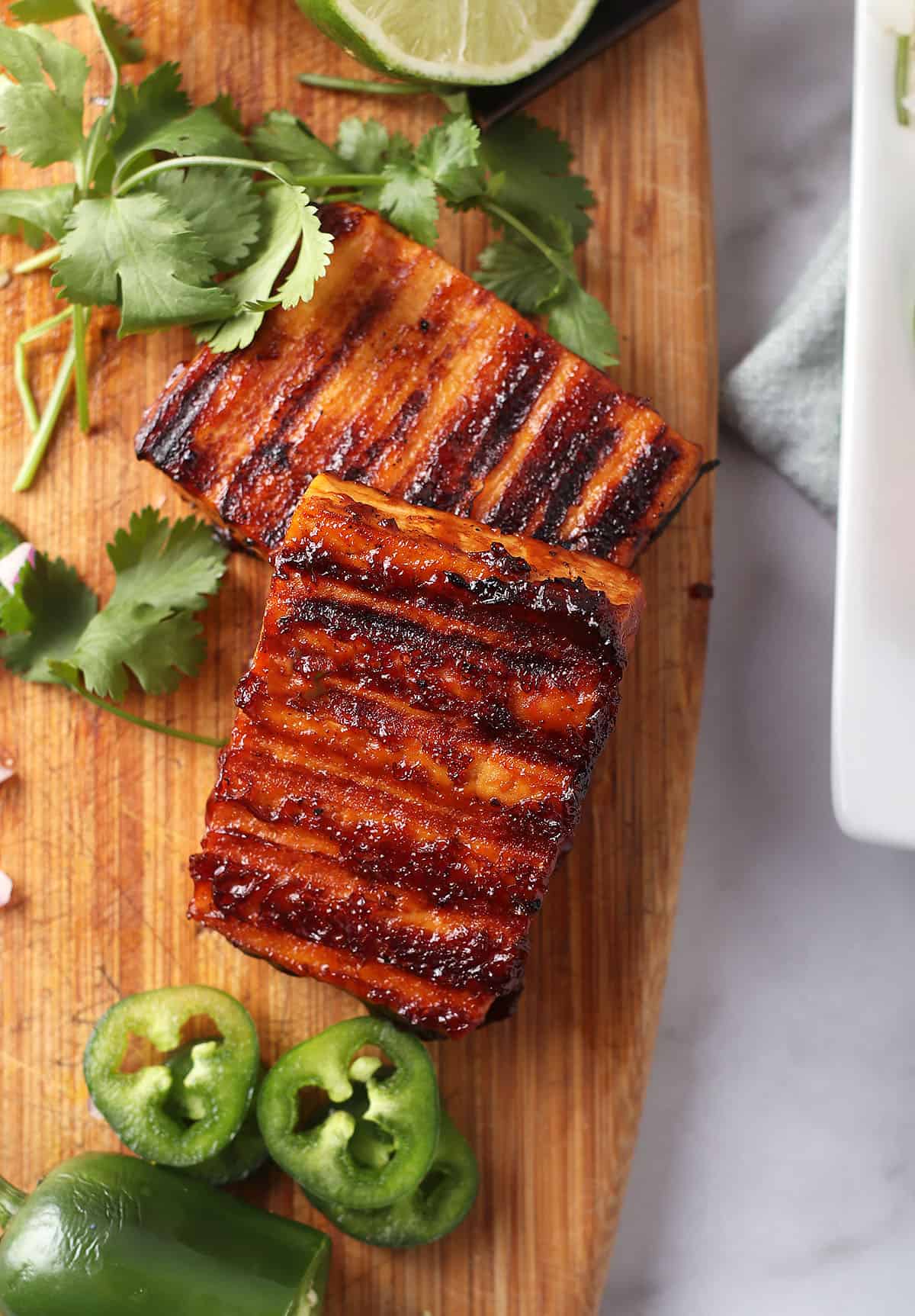 Two tofu steaks on a wooden cutting board, surrounded by sliced jalapenos and fresh cilantro.