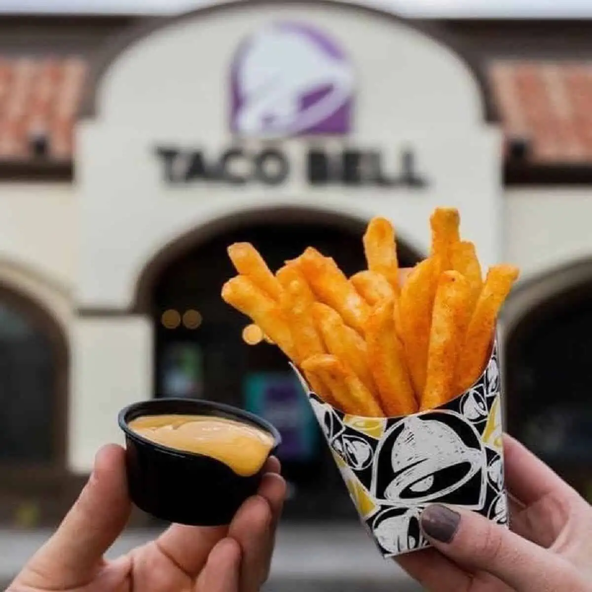 Two hands holding a black plastic cup of vegan nacho cheese and a cardboard container of fries held up in front of a Taco Bell restaurant.