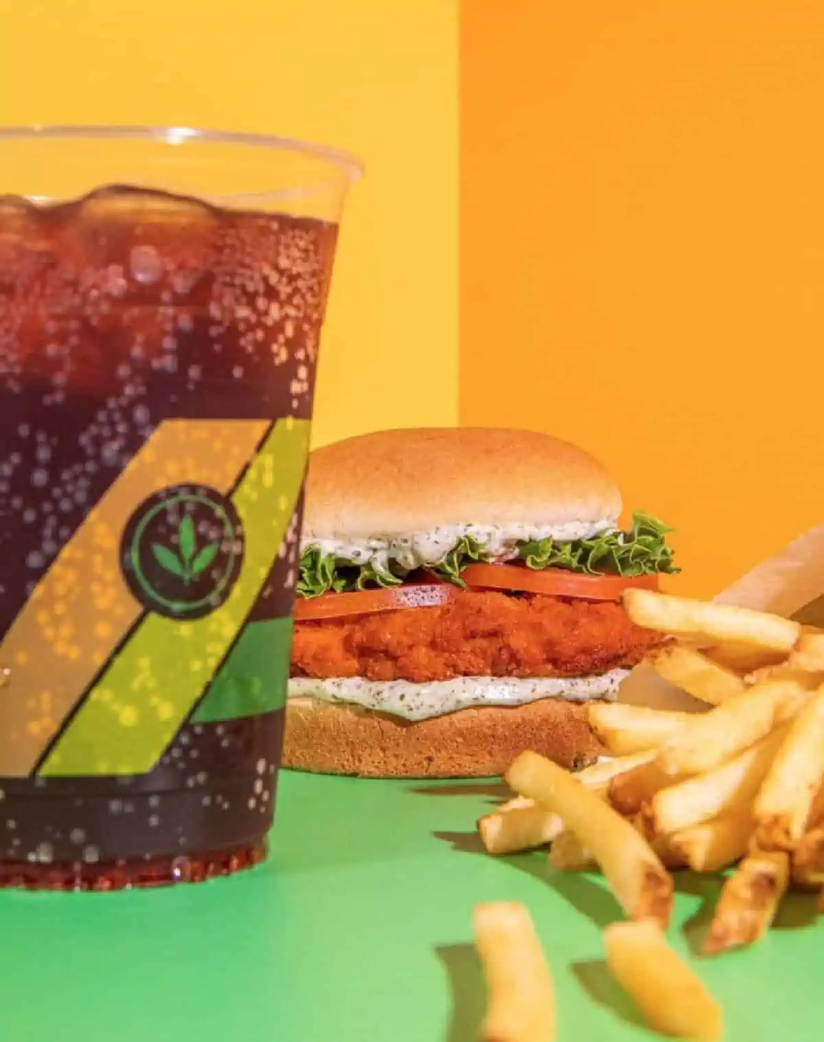 Plastic cup filled with cola, Plant Power Fast Food Buffalo 66 sandwich with fries on a green table with a background of yellow walls.
