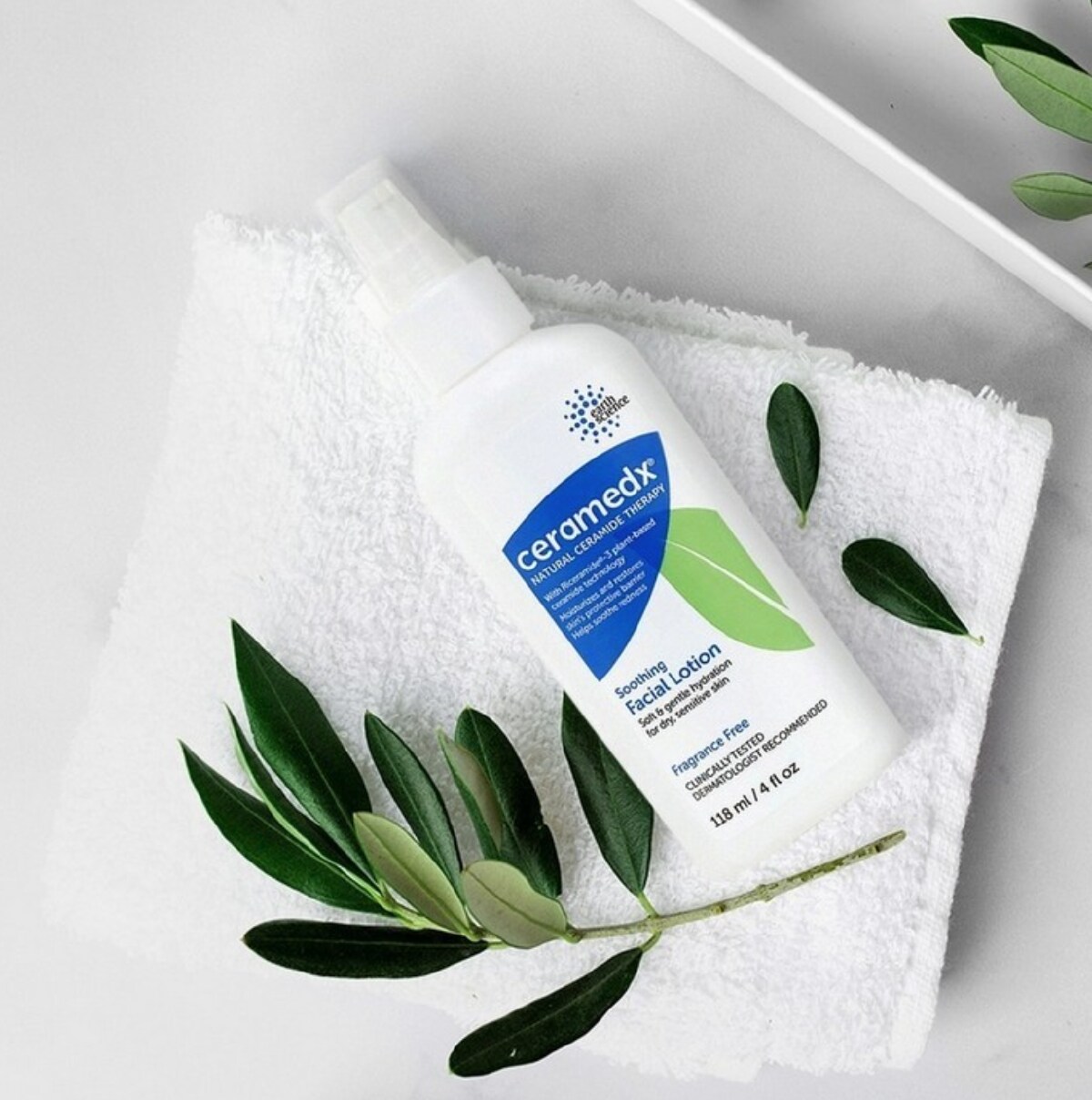 A white, blue and green bottle of Ceramedx face lotion on a white towel accented by a green plant cutting.