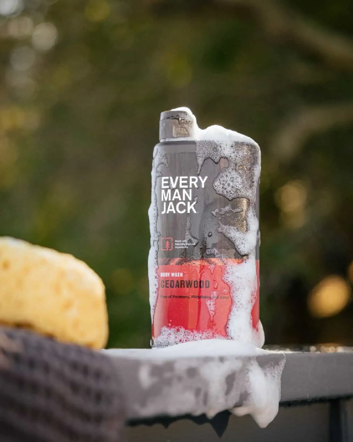 A red and brown, bubbly bottle of Cedarwood body wash by Every Man Jack against a nature background.
