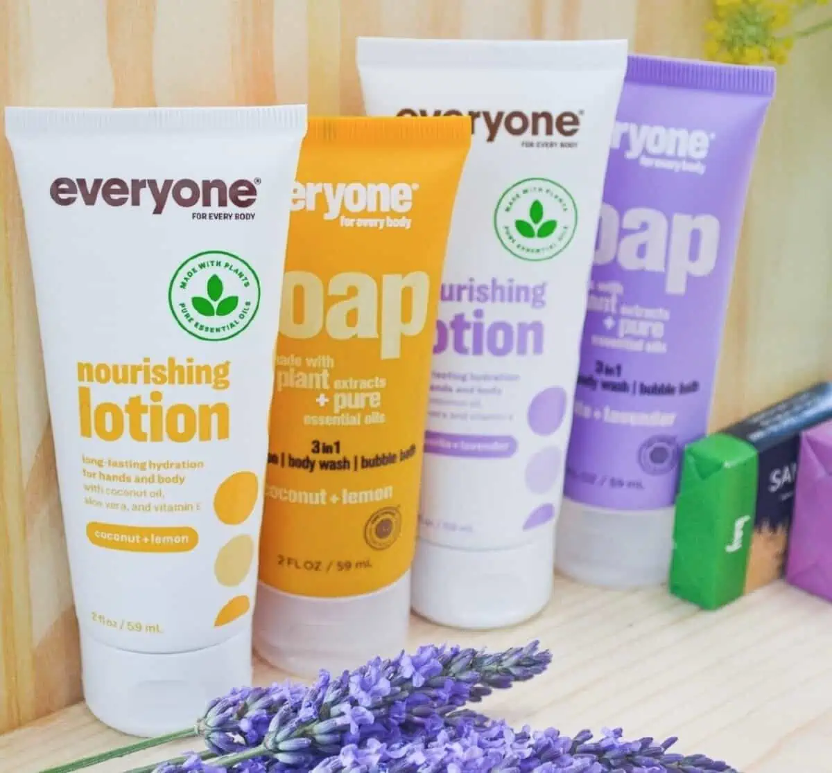 Four bottles of Everyone lotion and soap in white, orange, white, and purple bottles against pale wood paneling and accented with fresh lavender blossoms.