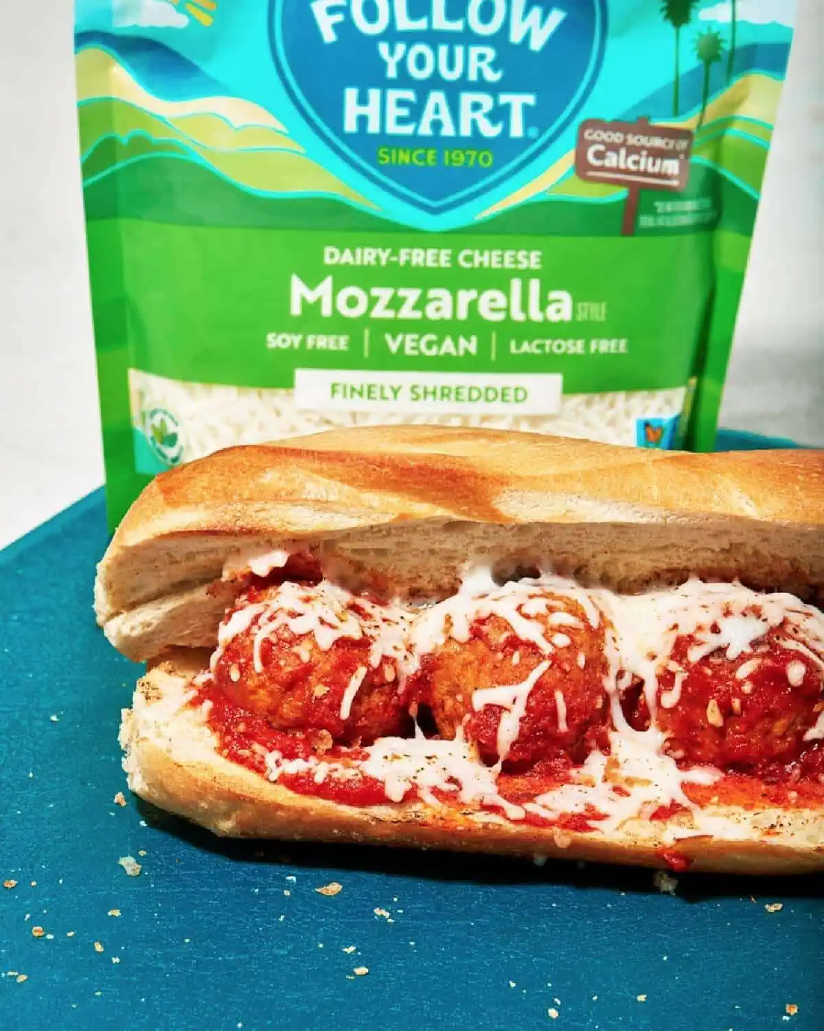 A green and blue pouch of Follow Your Heart mozzarella cheese and a vegan meatball sub on a blue countertop.