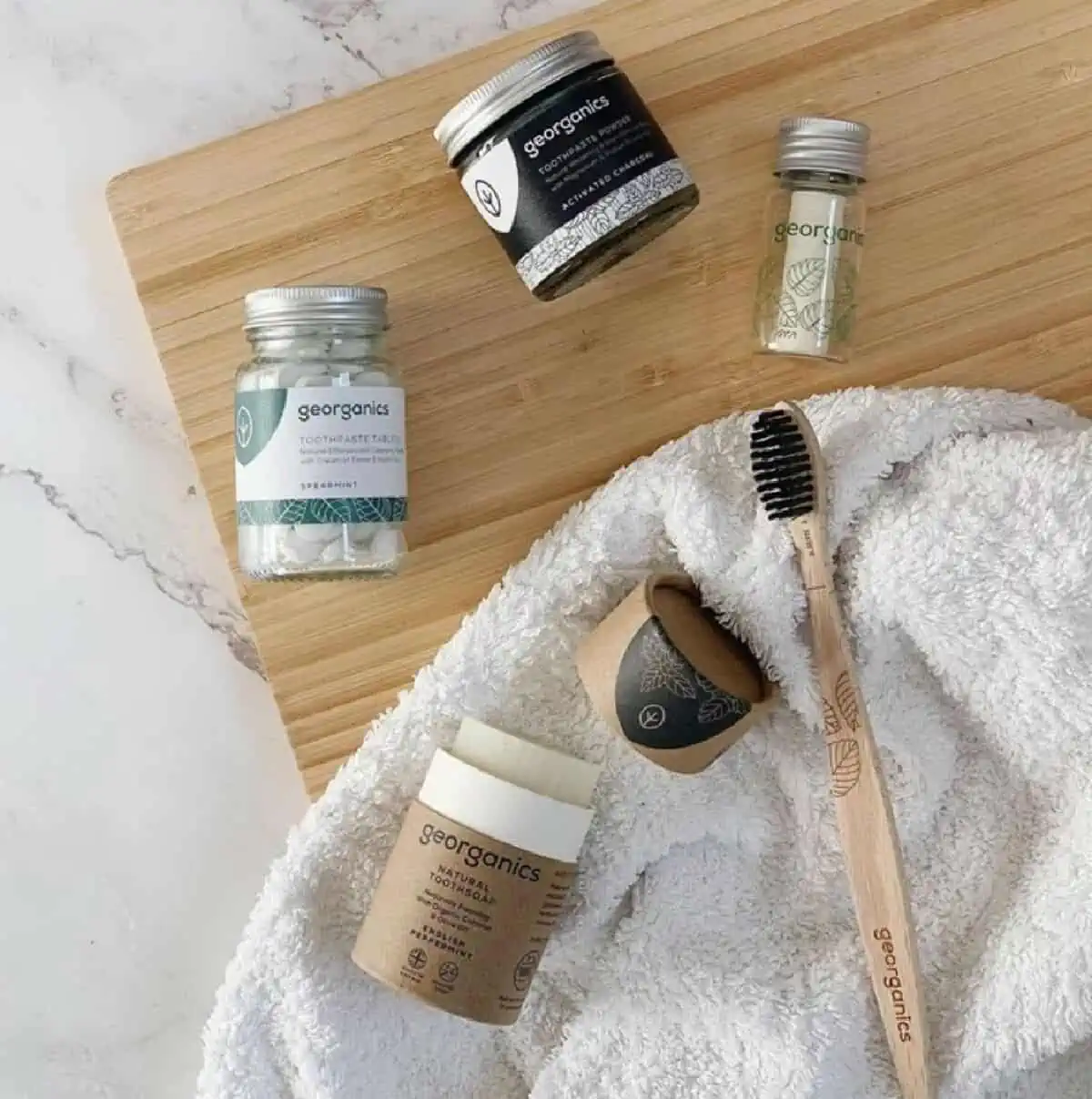 Three bottles of Georganics toothpaste tabs, mouthwash tabs and floss on a wooden cutting board next to toothpowder and a wooden tooth brush on a towel.