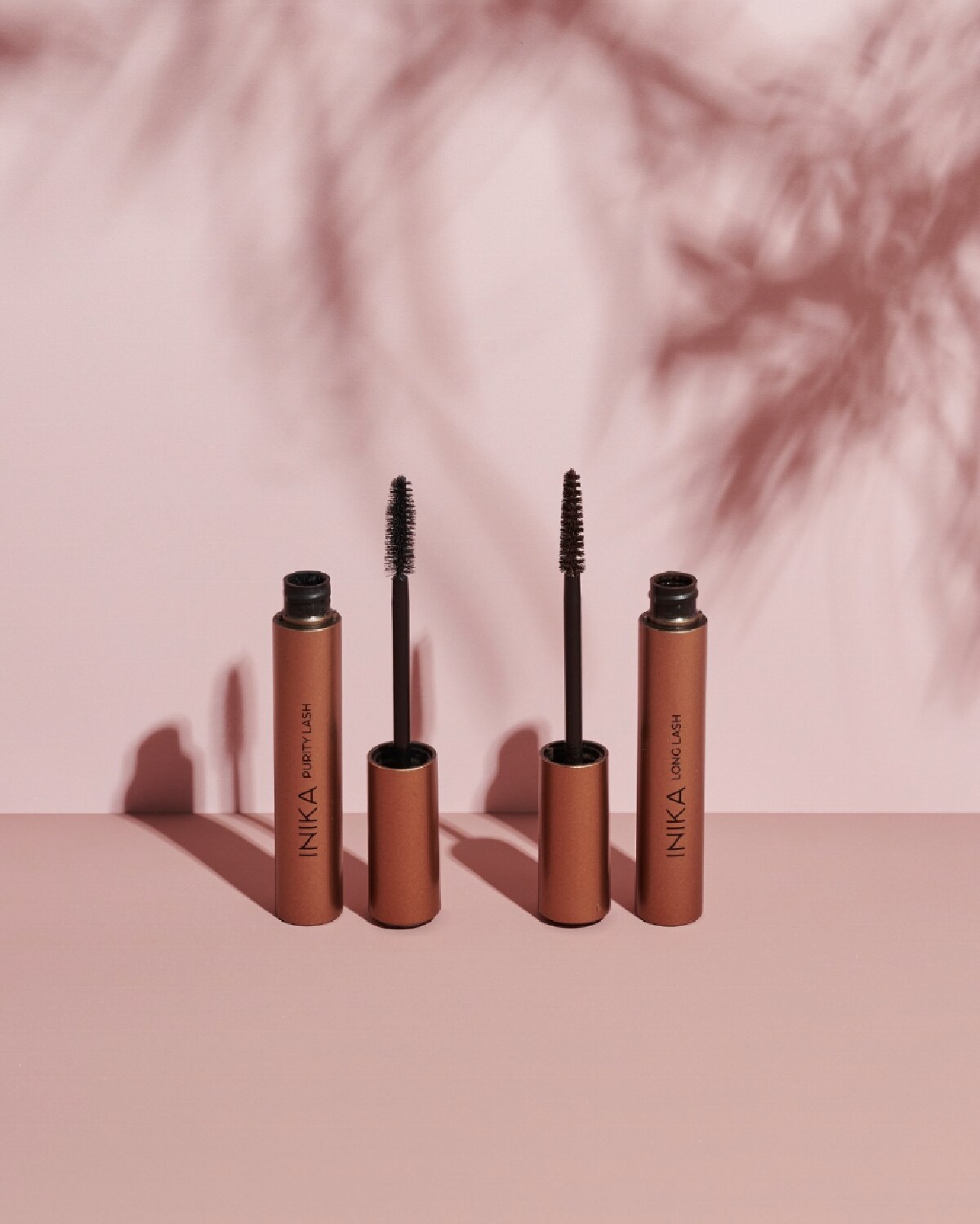 Two copper colored opened tubes of Inika Organics mascara next to black mascara wands against a shadowed pink backround.