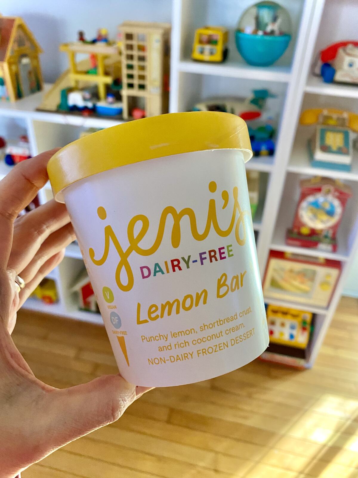 A hand holding a pint of Jeni's Dairy-Free Lemon Bar ice cream against a background of vintage Fisher Price toys.