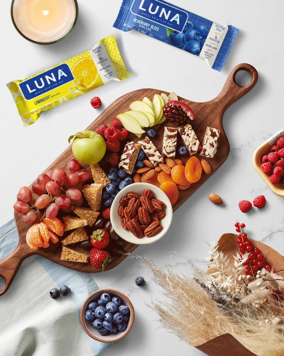 Two Luna vegan protein bars in Lemon and Blueberry next to a wooden cutting board filled with fruit and nuts.