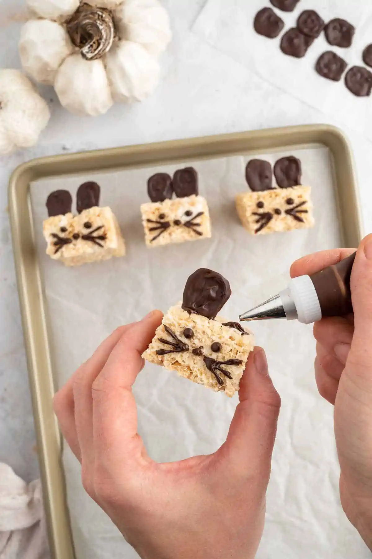 A hand hold a mice krispy while it pipes chocolate onto its head to attach the ears.