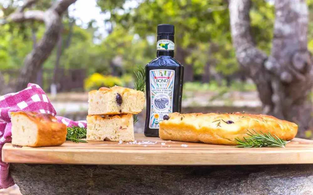 A wooden cutting board holding a bottle of Bono Olive Oil, rosemary vegan focaccia bread, a red and white checked napkin and fresh sprigs of rosemary.
