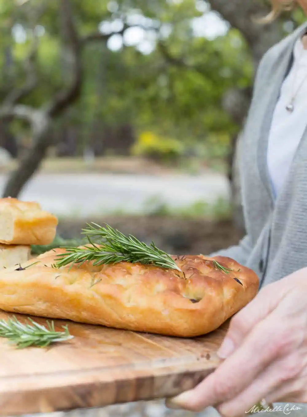 A woman in a gray sweater and white shirt is holding a wooden board with a large loaf of focaccia bread on it topped with fresh rosemary.