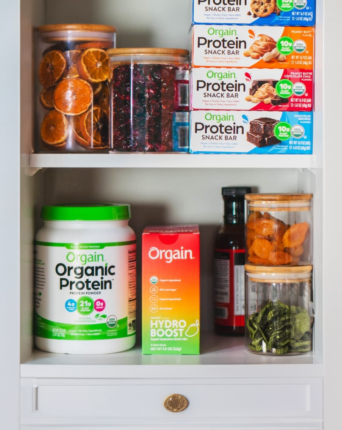 A cupboard filled with Orgain protein snack bars and protein powder, surrounded by jars of dried fruits and packets of hydroboost.