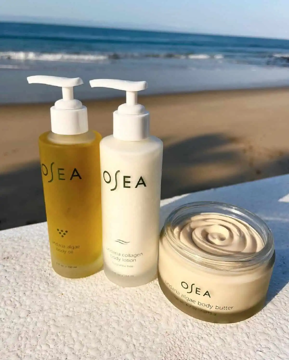 Two glass bottles of Osea oil and lotion with white pump tops and standing next to a jar of cream in front of a beach setting.
