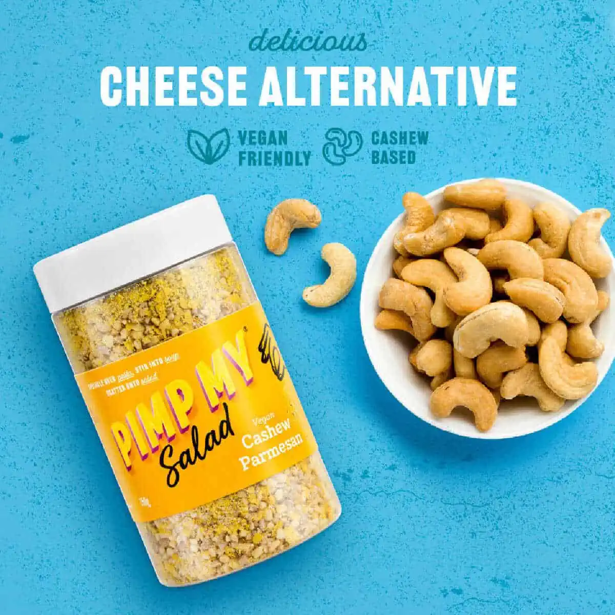 A clear and yellow plastic container of Pimp My Salad cashew vegan parmesan with a white cap next to a white bowl of cashews on a blue background.