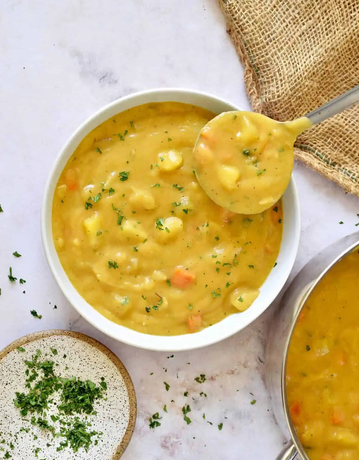 Ladle of vegan potato soup in a large bowl next to a pot of creamy soup.