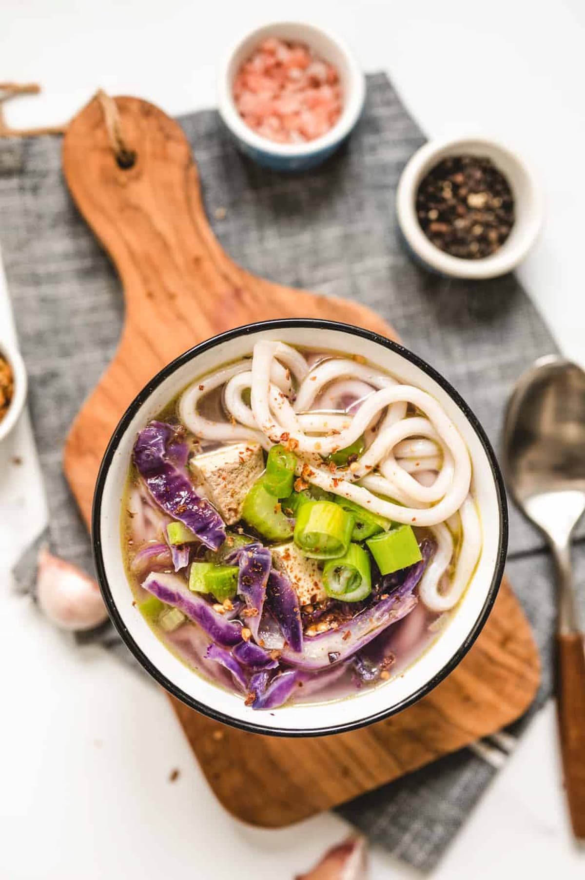 Bowl of purple vegan udon noodle soup with cabbage, green onions, tofu, and thick noodles, served on a wooden board with salt and pepper bowls in the background.