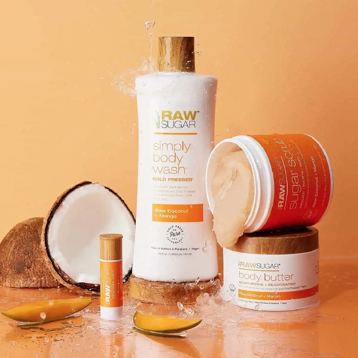 A white bottle of Raw Sugar Simple Body Wash next to two containers of body scrub, body butter, a lip balm and slices of coconut and mango against an orange background.