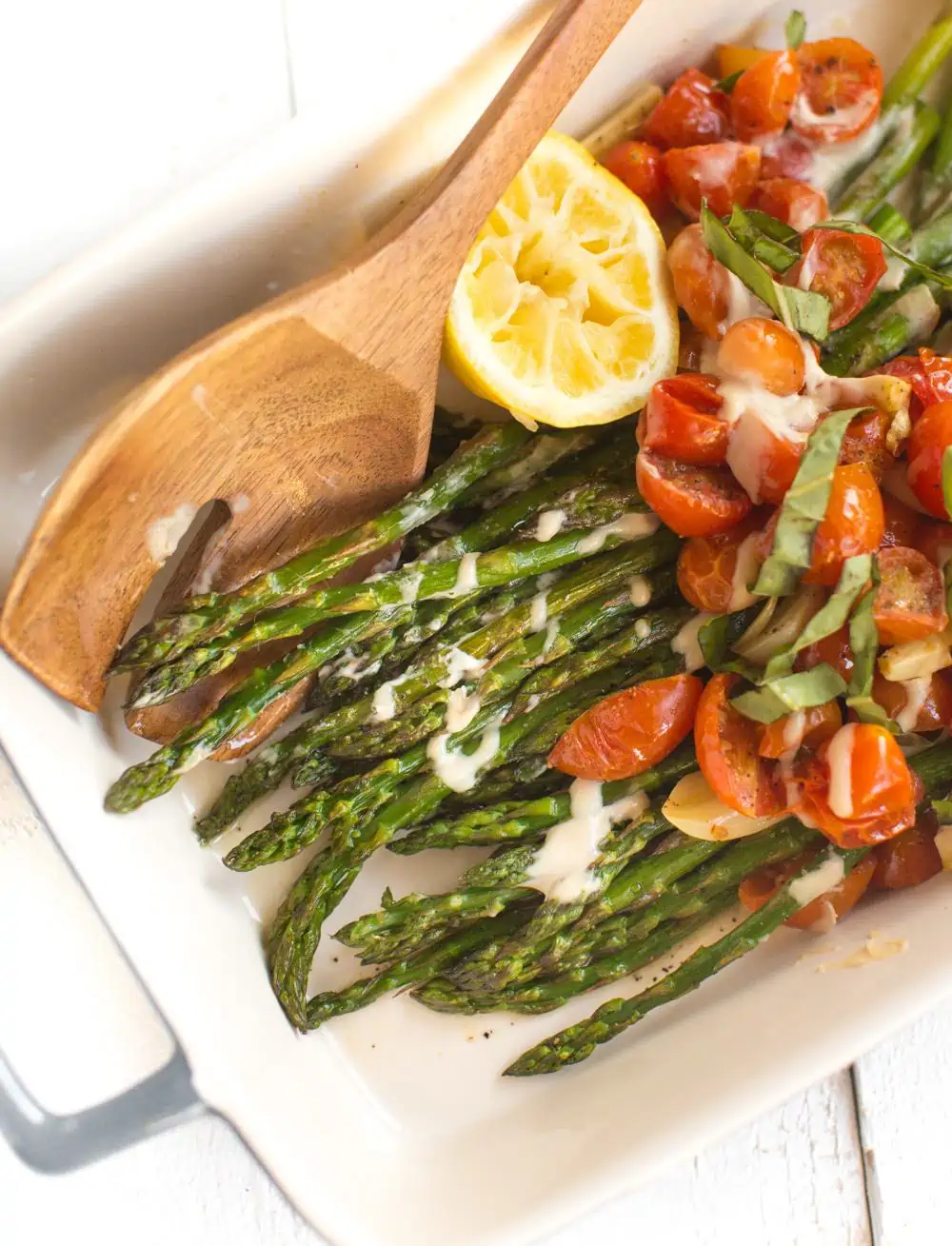 Roasted asparagus and tomatoes with tahini drizzle in a serving dish with a wooden spoon.