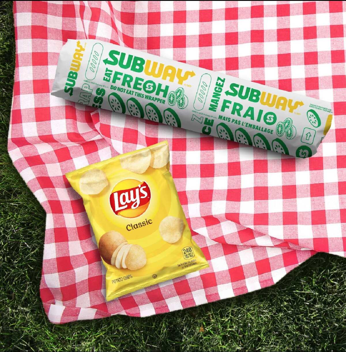A wrapped Subway sandwich and a bag of Lay's potato chips laid out on red gingham blanket on the grass.