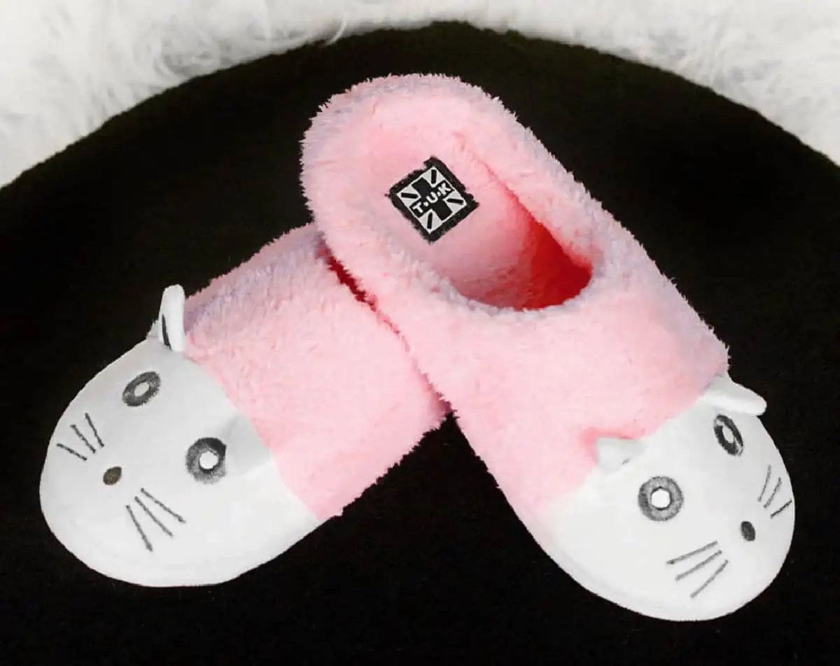 An adorable pink and white pair of kitty slippers crossed at the heel on the black background.