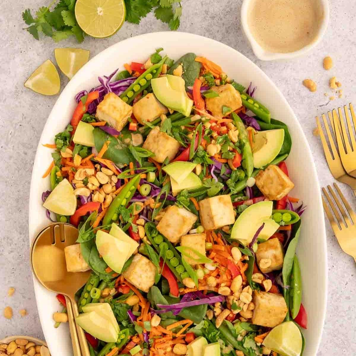 A large white bowl filled with Thai tofu salad and surrounded by lime wedges, peanut butter sauce and gold forks.