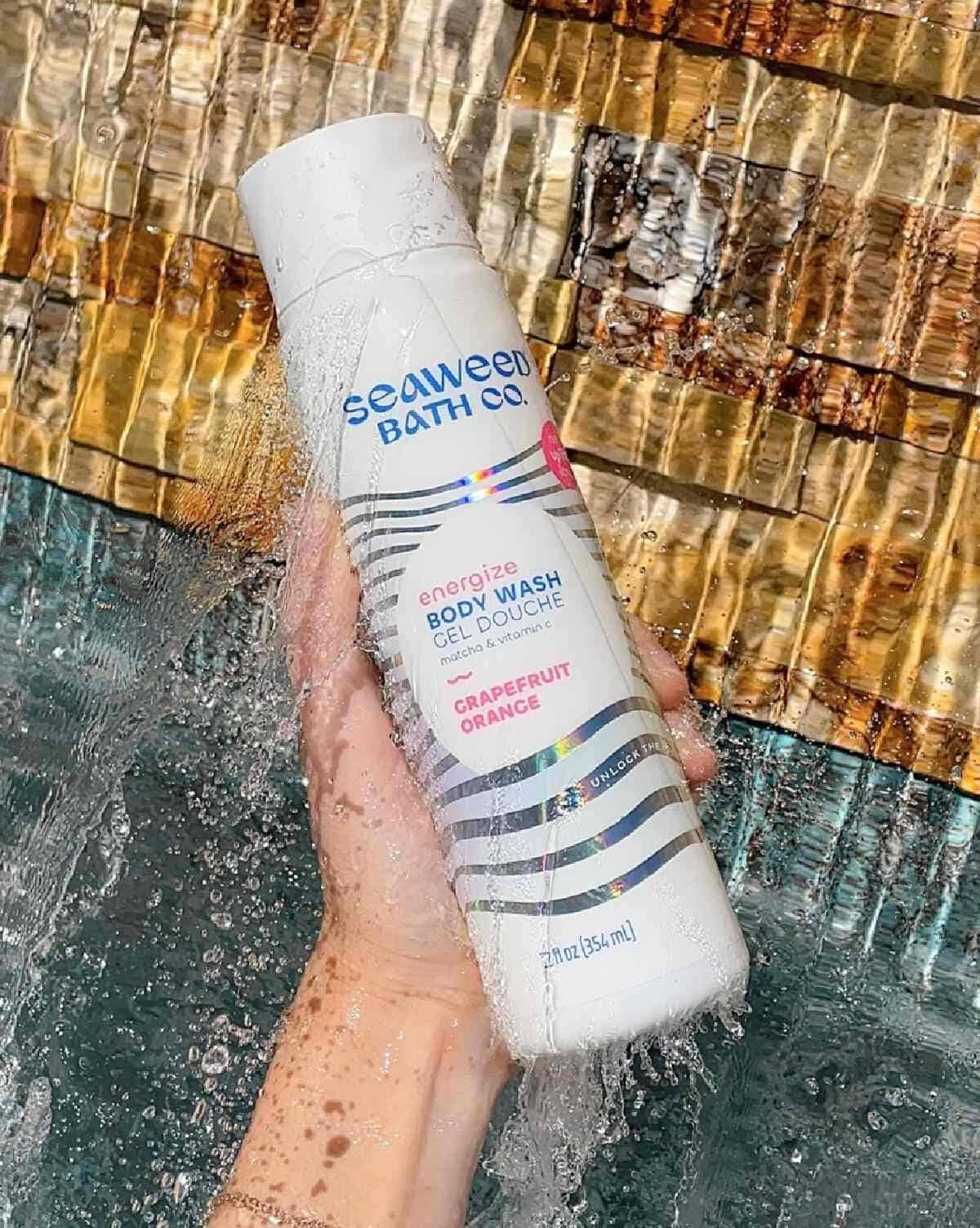 A hand holding a white bottle of Energize Body Wash by Seaweed Bath Co. against a watery brown and blue background.