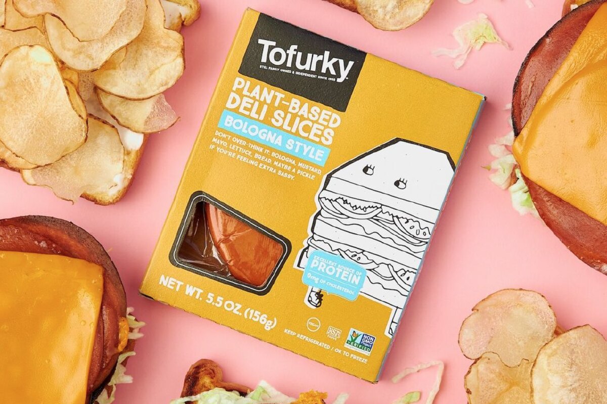 Tofurky Plant-based deli bologna slices in a mustard yellow colored box next to potato chips and vegan bologna sandwiches against a pink background.