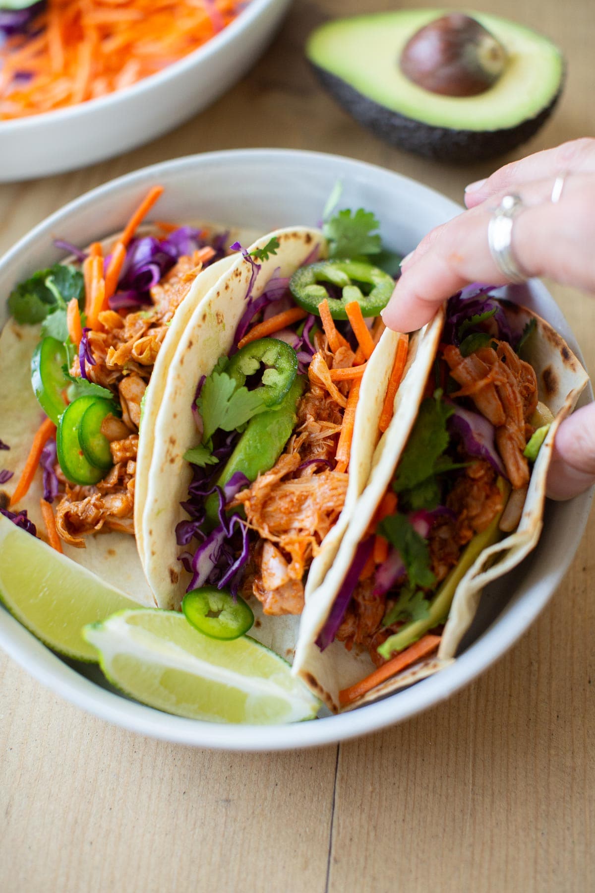A hand holding one of three vegan jackfruit tacos with barbecue sauce from a white plate with lime wedges.