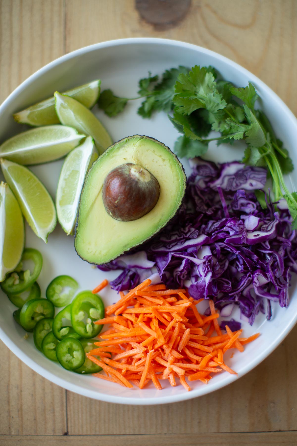 A white plate holding the toppings for vegan bbq jackfruit tacos including: avocado, sliced red cabbage, shredded carrots, cilantro, lime wedges and jalapeno peppers.