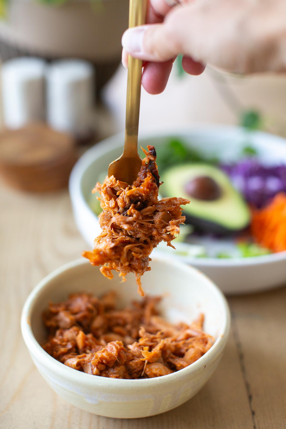 A hand holding a fork full of homemade bbq jackfruit to add to vegan tacos. A white bowl of veggie toppings is in the background.