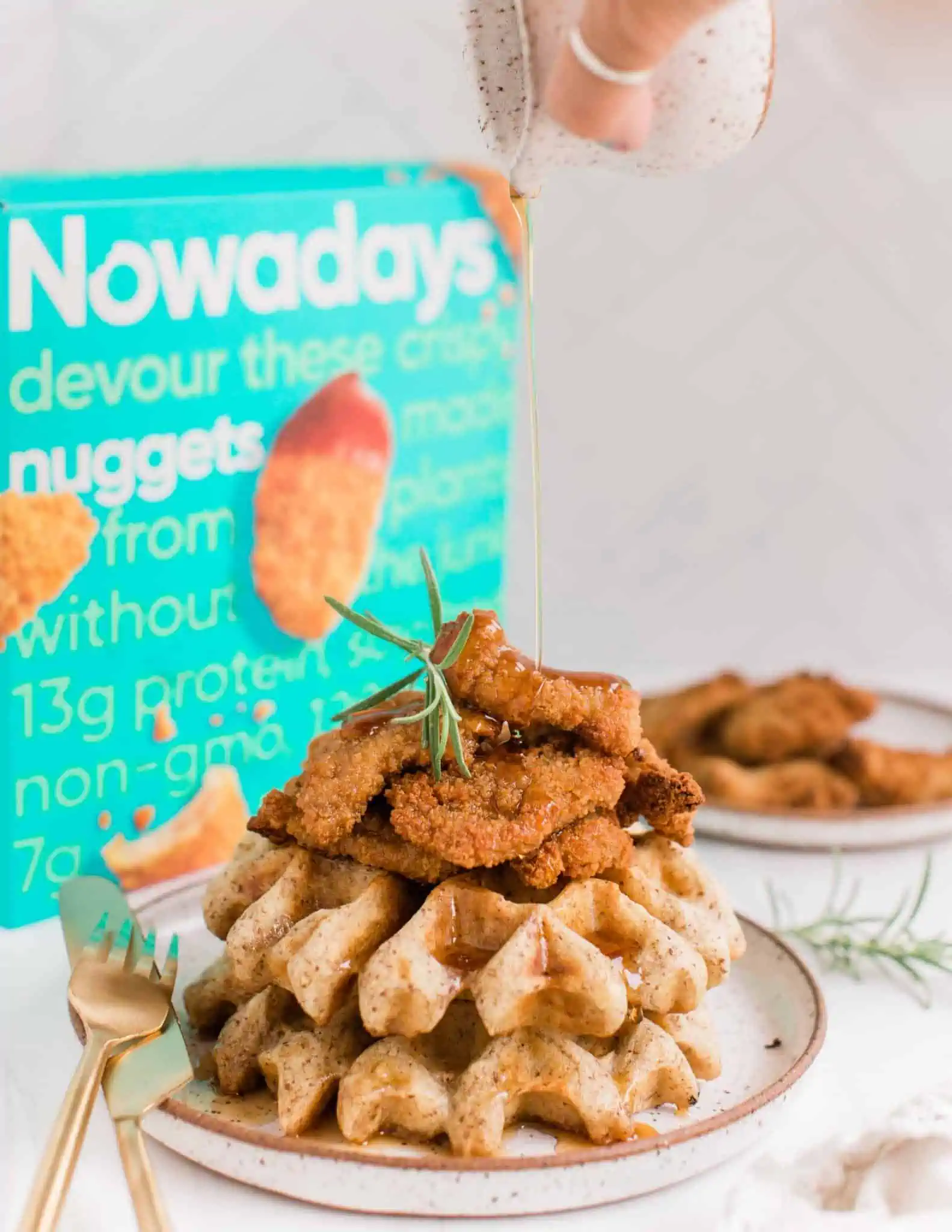 Vegan Chicken and Waffles Stacked on a Plate With a Hand Pouring Maple Syrup on Top in front of a Nowadays Vegan Nuggets Box.
