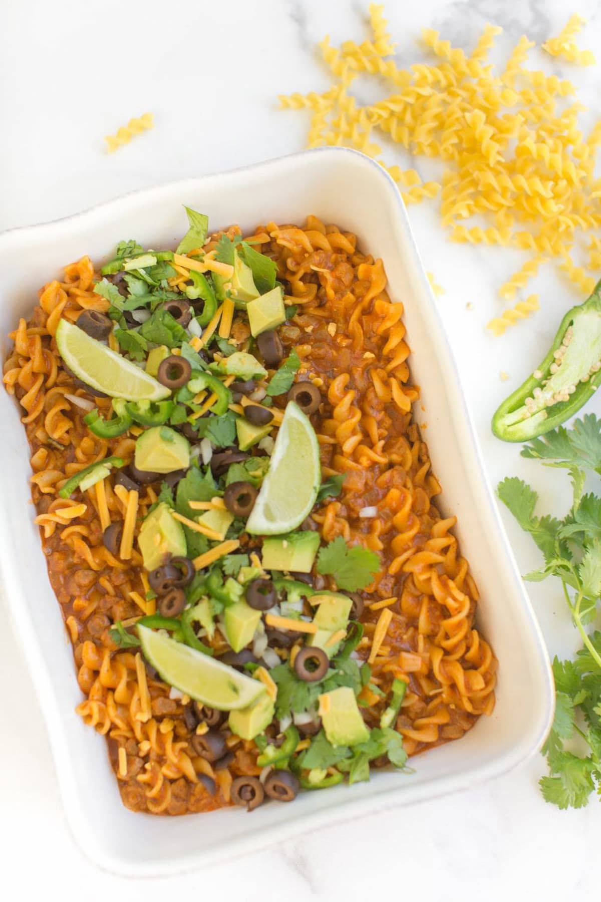 White rectangular baking dish of vegan enchilada pasta garnished with cubed avocado and lime on a white countertop.