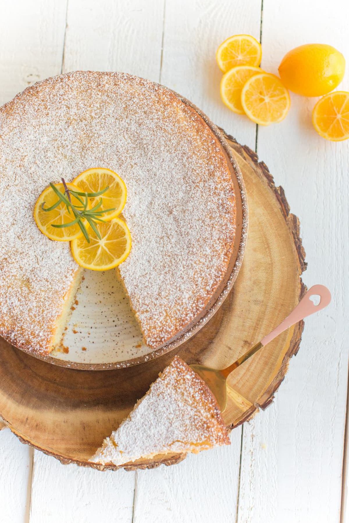 A vegan lemon olive oil cake on a round wooden cutting board with a slice cut out and garnished with lemon slices.