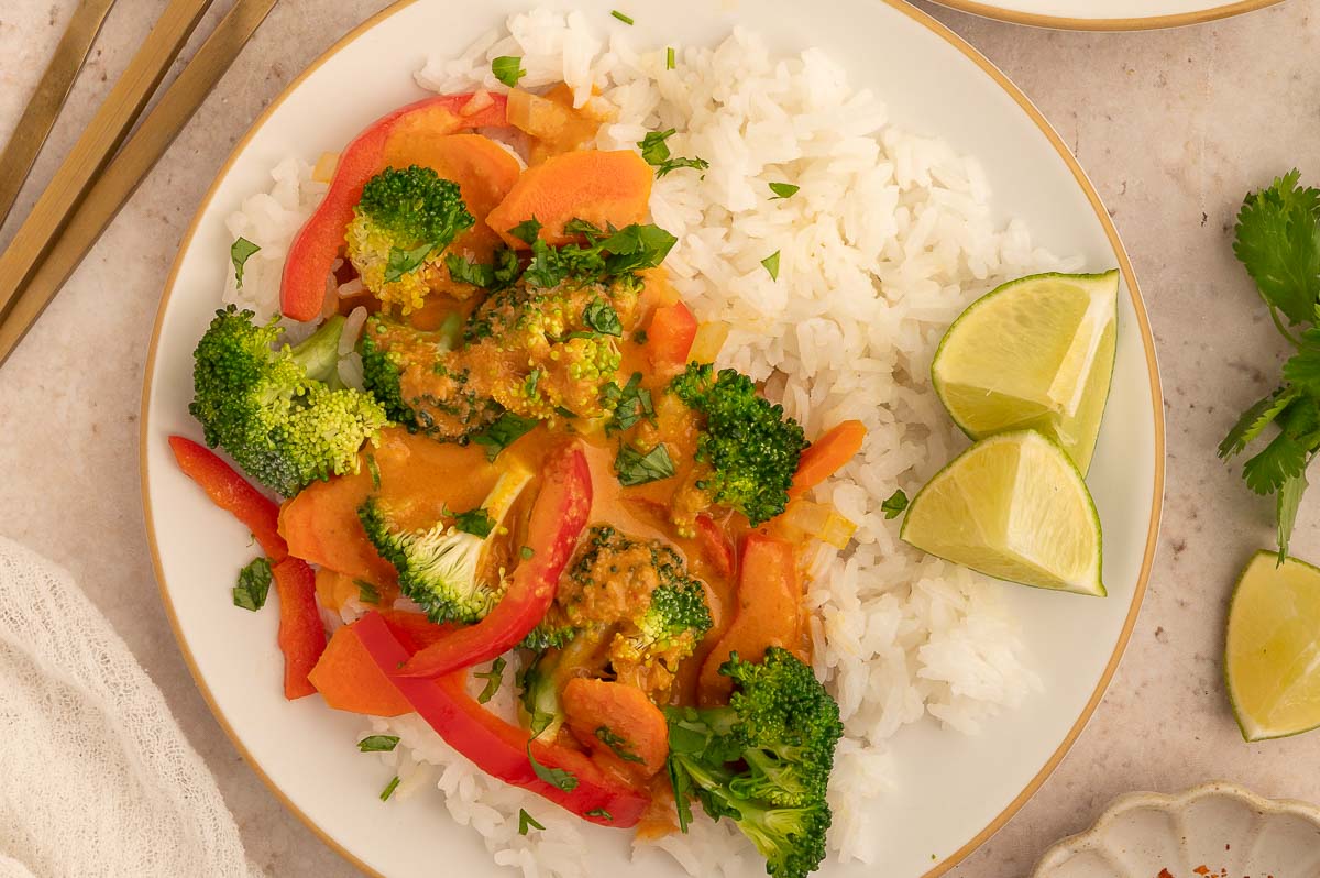 Vegan panang curry with white rice, broccoli, bell peppers, and lime wedges on a white plate.