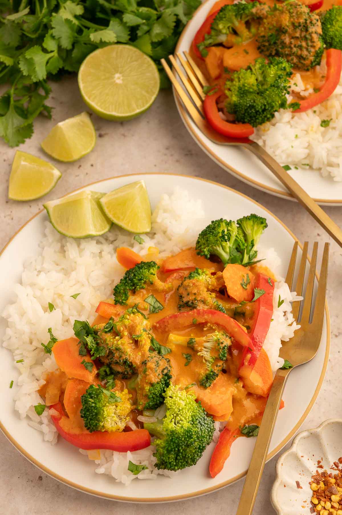 Vegetarian panang curry with white rice, broccoli, bell peppers, and lime wedges on a white plate with a gold fork and next to a second plate of food.