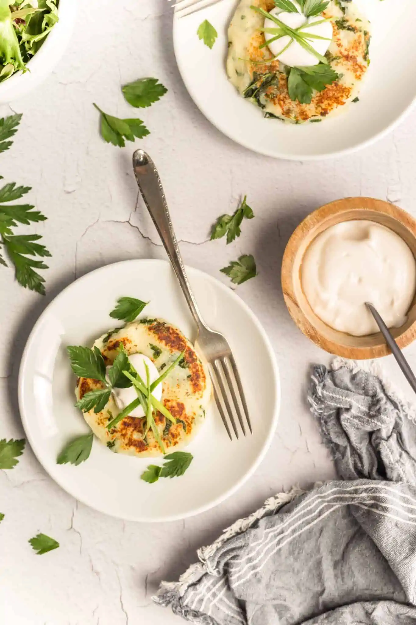 Two white plates of vegan potato cakes Recipe with kale topped with chopped fresh parsley and accompanied by a small bowl of vegan mayo. A gray napkin is on the bottom of the photo.