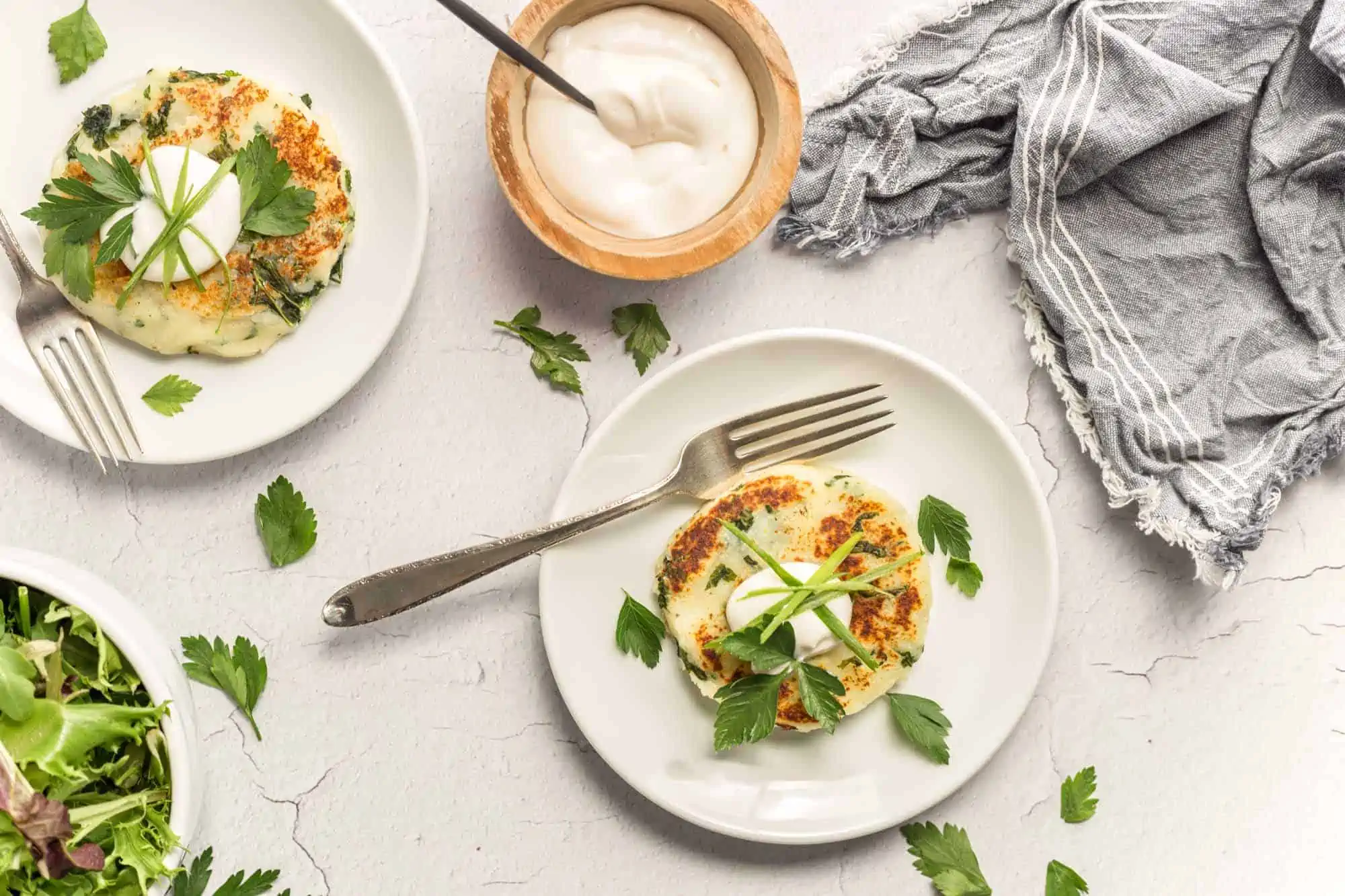 Individual vegan potato cakes on white plates on a white table along with sprinkled herbs, a gray napkin and a small bowl of vegan mayo.