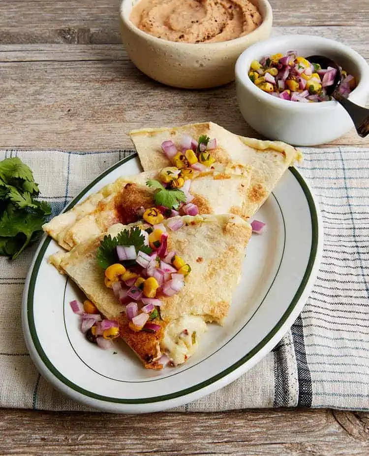 Three vegan quesadillas on a plate topped with diced red onion, corn kernels, and cilantro leaves.