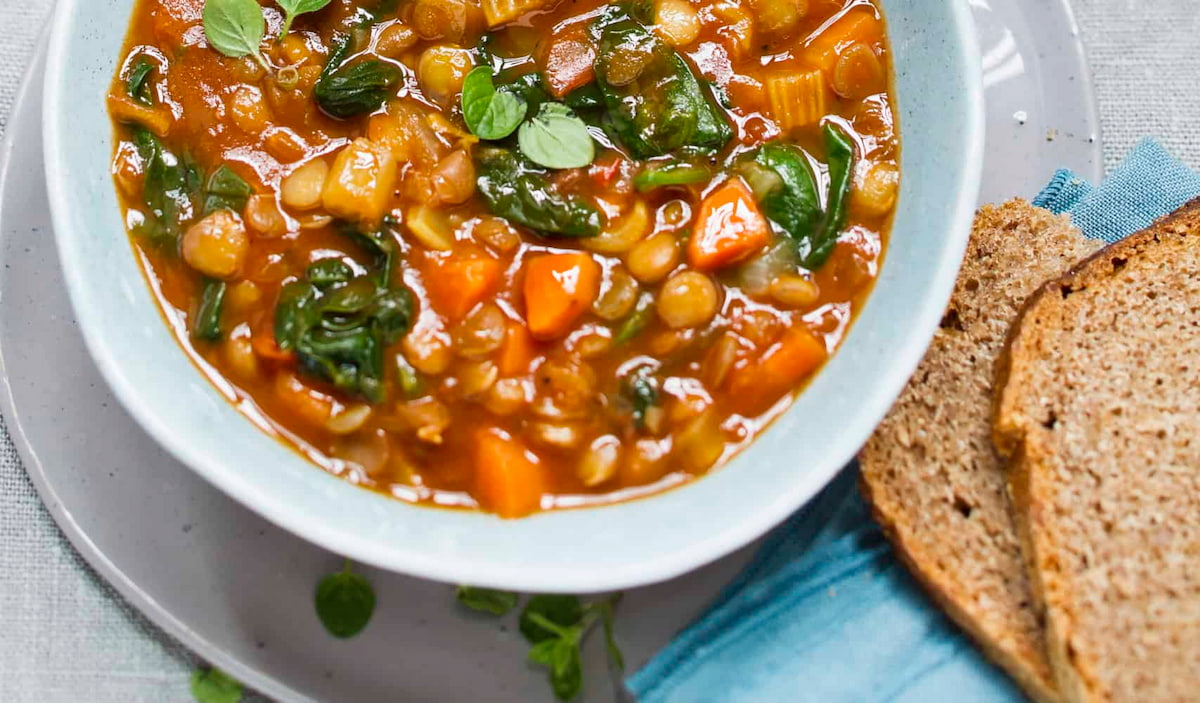 A bowl of hearty crockpot tomato lentil stew with veggies in a white bowl next to a slice of whole grain bread.