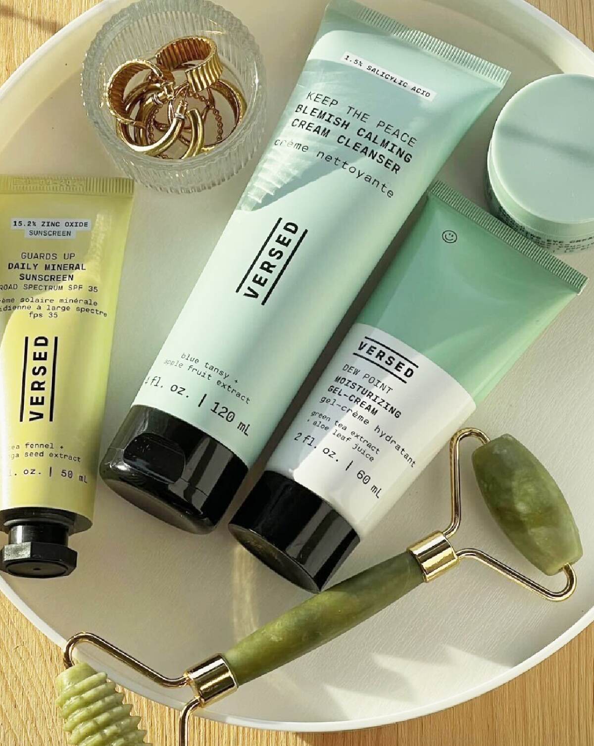 A white dish including three yellow or green tubes of Versed skincare products next to a jade roller and a dish of gold rings.