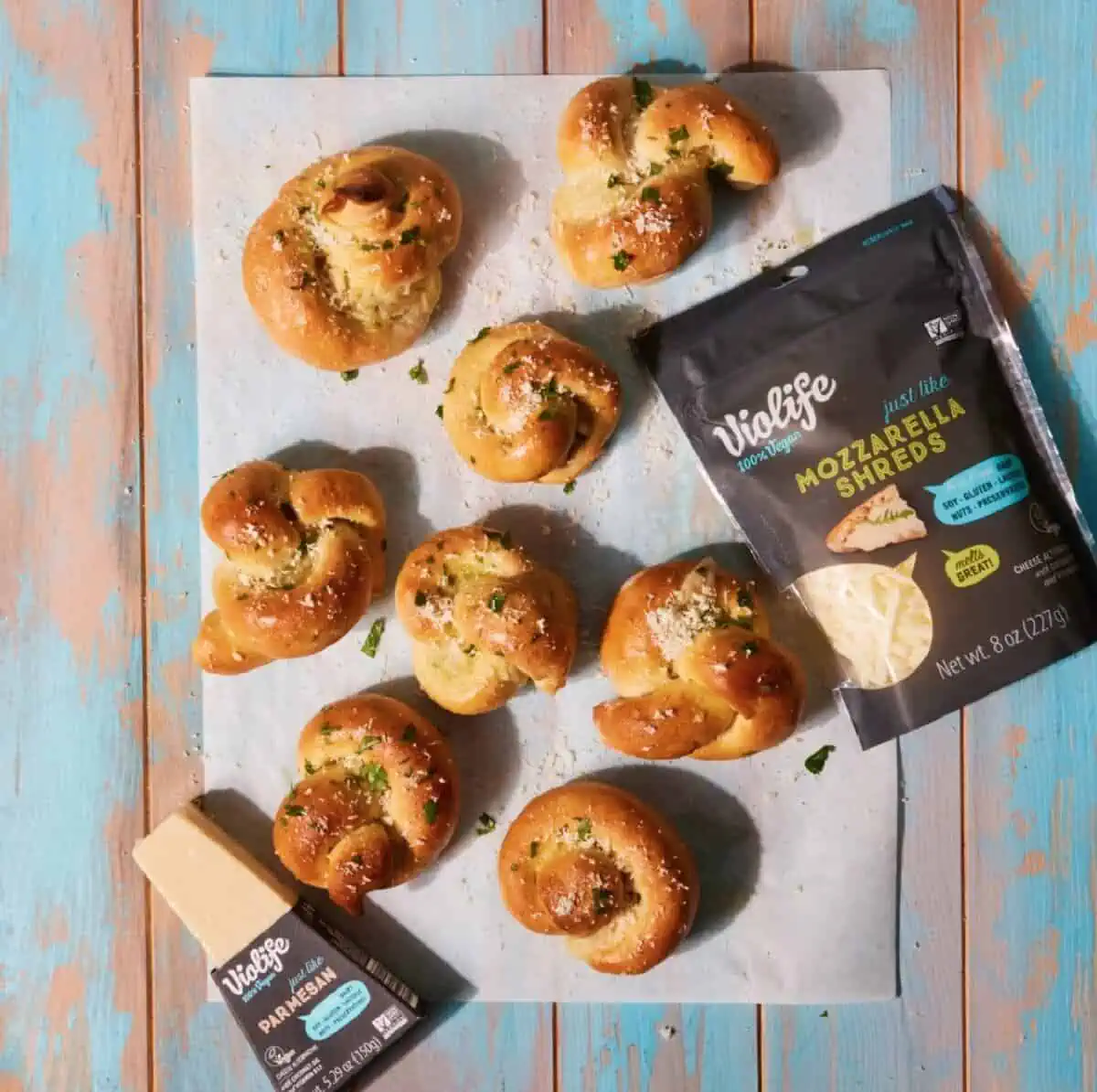 Eight pretzel twists on a piece of parchment against a wooden floor along with Violife Parmesan and Mozzarella cheese.