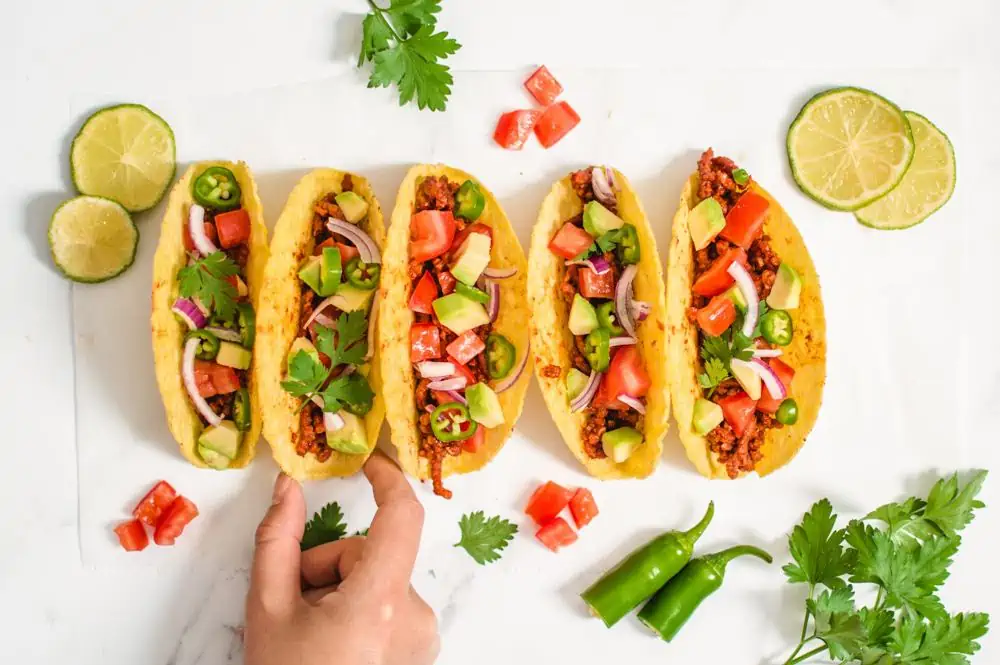 A hand reaching for a vegan taco in a line up of five crunchy corn tacos on a piece of white parchment paper.