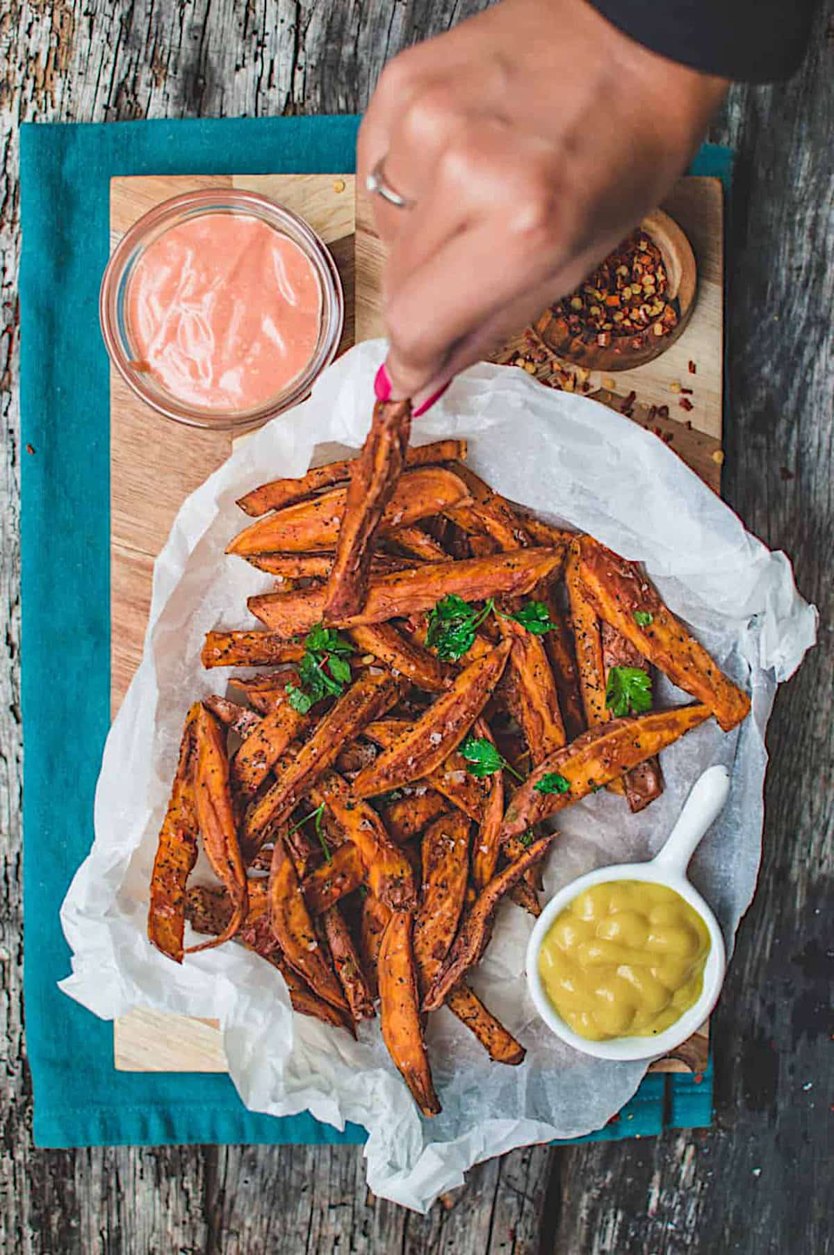 A hand reaching for an air fried sweet potato fry from a pile of fries, with two different dips served on the side.