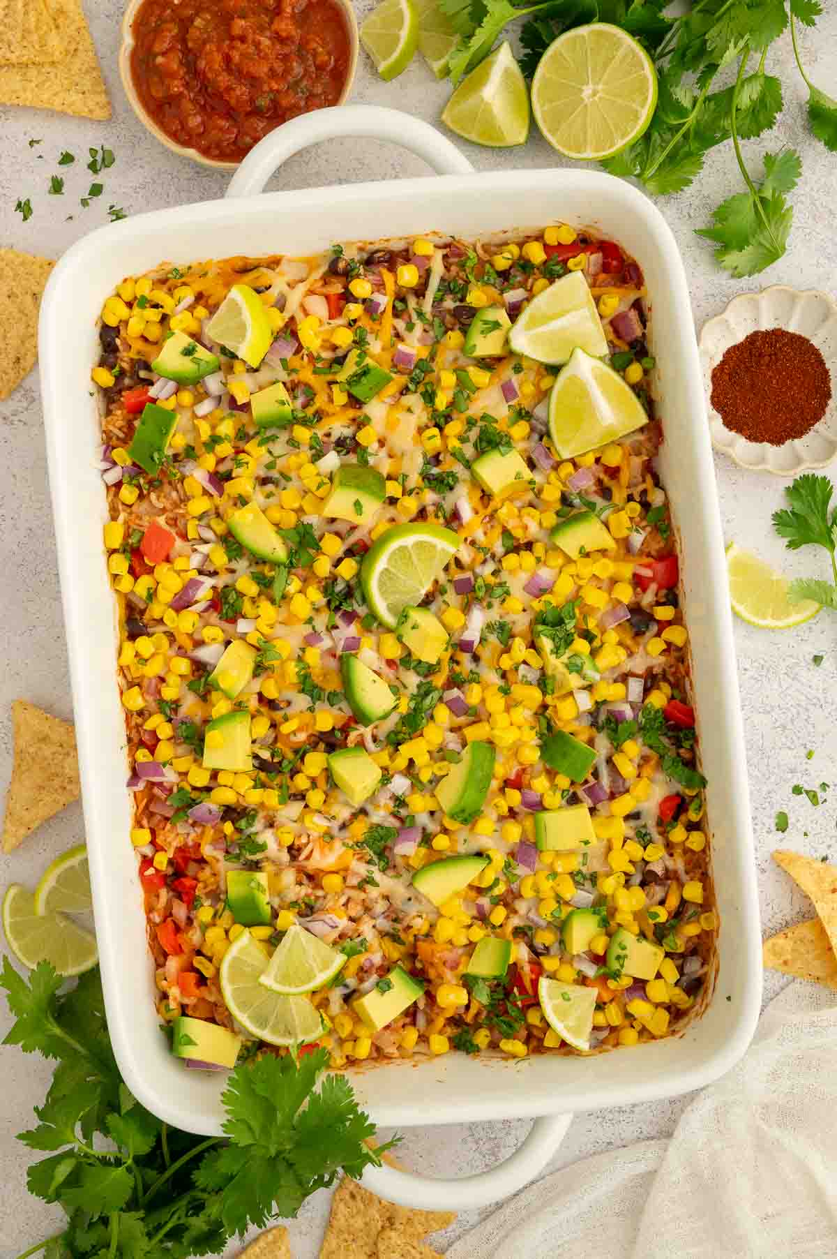 A large casserole dish of black bean and rice casserole topped with Mexican-style toppings like cilantro, red onion, cilantro, avocado, corn, and lime slices.