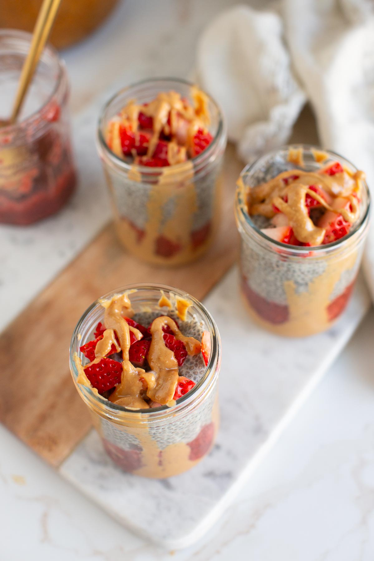 Three chia pudding parfaits with strawberry jam and peanut butter drizzle in glass jars.