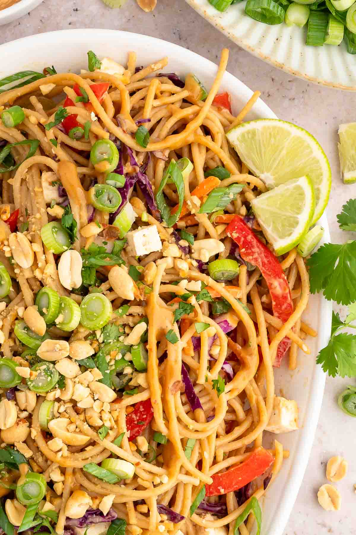 A large serving platter of cold noodle salad topped with roasted peanuts, sliced green onions, and lime wedges.