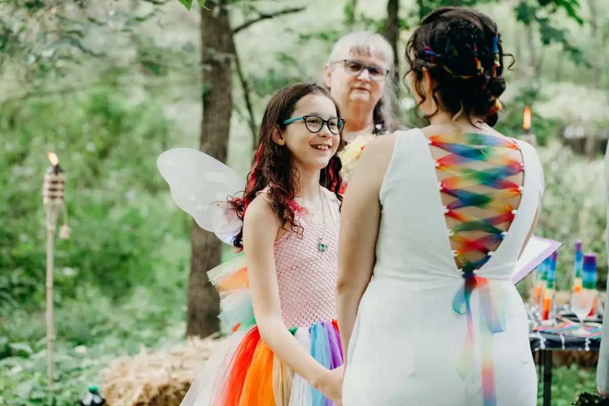 Bride reading vows to her stepdaughter during rainbow wedding in a forest setting.