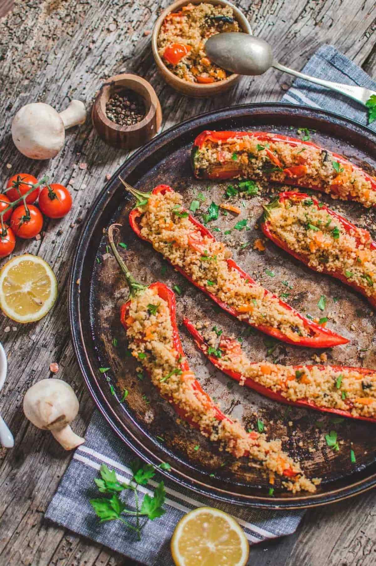 Roasted couscous stuffed peppers on a tray with fresh tomatoes, lemon, and mushrooms on the side.