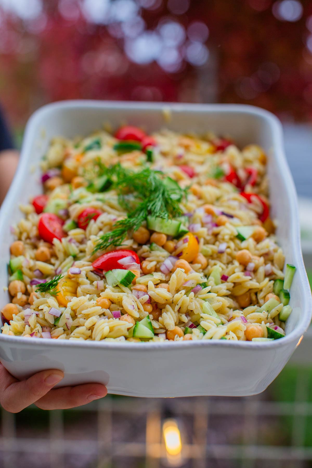 Hands holding up a large serving dish filled with chickpea orzo salad topped with fresh dill.